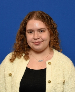 woman with medium blond curly hair in a yellow cardigan and black top smiles; blue background