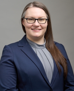 woman with medium brown hair, wearing glasses, an gray top and a blue blazer smiles