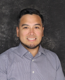 Portrait of a smiling man with short dark hair and a trimmed beard, wearing a gray checkered button-down shirt against a dark textured background.