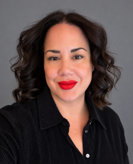 Professional headshot of a smiling woman with shoulder-length, dark brown wavy hair, wearing a black textured collared shirt and bright red lipstick. She faces the camera against a neutral gray studio background.