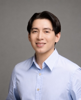 Professional headshot of a smiling man with dark, side-parted hair wearing a light blue dress shirt against a plain gray background.