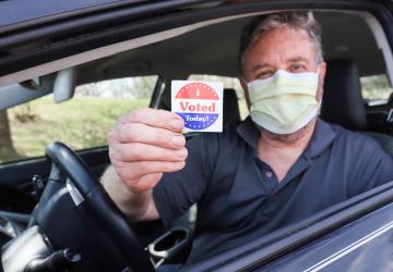 Man wearing a surgical mask for COVID-19 pandemic holds a sticker that says "I VOTED"