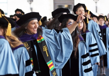 A student wearing academic regalia waves from the processional at the 2017 Graduation.