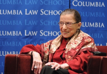 Ruth Bader Ginsburg smiling while wearing a red suit in front of a blue Columbia backdrop.