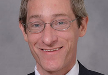 Headshot of a man wearing glasses, a white shirt, black blazer, and red tie