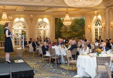 Person on a stage speaking to audience members seated at tables in a dining room