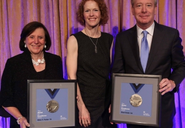 A woman in black flanked by a woman and man, also in black, both holding framed medals.