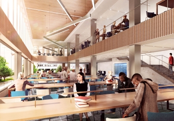 Students sit at long tables next to a bank of windows and beneath a mezzanine.
