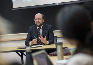 Man in glasses and suit at table in front of a classroom