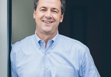 Dark haired man in button down shirt leaning against doorframe and smiling.