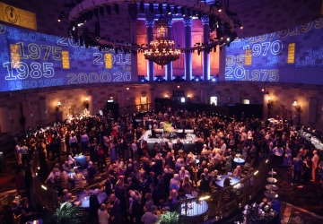 Wide shot of room filled with people at a cocktail party with Columbia logo projections on the walls. 