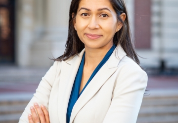 Elora Mukherjee in white jacket and blue blouse with arms crossedd in front of a Columbia University building