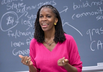Olatunde Johnson in bright pink dress standing before blackboard covered in writing