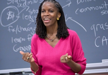 Olatunde Johnson in bright pink dress standing before blackboard covered in writing