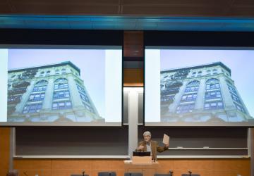 Professor Vincent Blasi gestures in front of a screen featuring a multistory building.