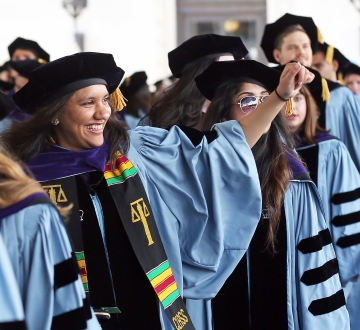 A student wearing academic regalia waves from the processional at the 2017 Graduation.