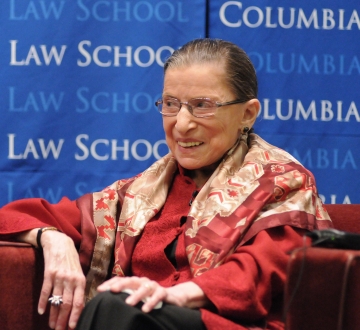 Ruth Bader Ginsburg smiling while wearing a red suit in front of a blue Columbia backdrop.