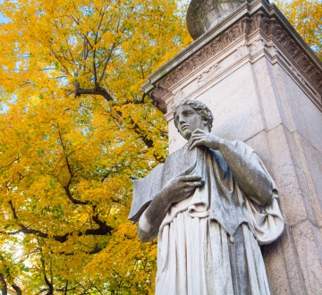 A statue of a figure holding a book in front of a tree with yellow leaves
