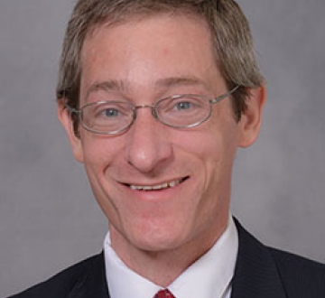 Headshot of a man wearing glasses, a white shirt, black blazer, and red tie