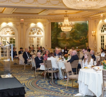 Person on a stage speaking to audience members seated at tables in a dining room