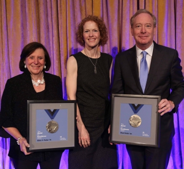 A woman in black flanked by a woman and man, also in black, both holding framed medals.