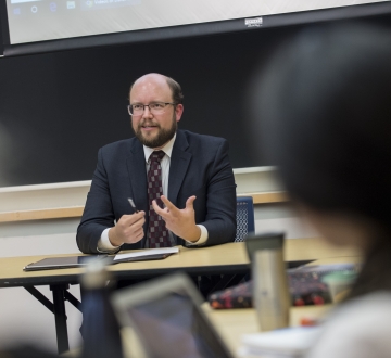 Man in glasses and suit at table in front of a classroom