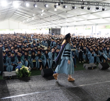 Woman in academic regalia crosses stage before large audience under enormous tent