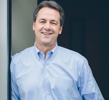 Dark haired man in button down shirt leaning against doorframe and smiling.