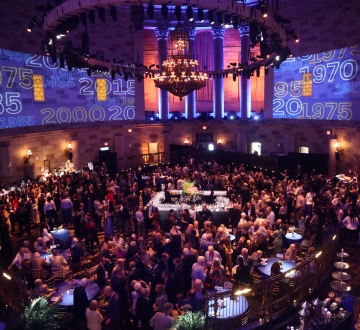 Wide shot of room filled with people at a cocktail party with Columbia logo projections on the walls. 