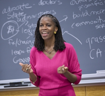 Olatunde Johnson in bright pink dress standing before blackboard covered in writing