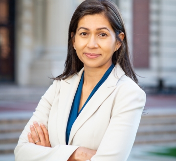 Elora Mukherjee in white jacket and blue blouse with arms crossedd in front of a Columbia University building