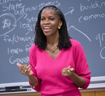 Olatunde Johnson in bright pink dress standing before blackboard covered in writing
