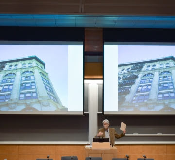 Professor Vincent Blasi gestures in front of a screen featuring a multistory building.