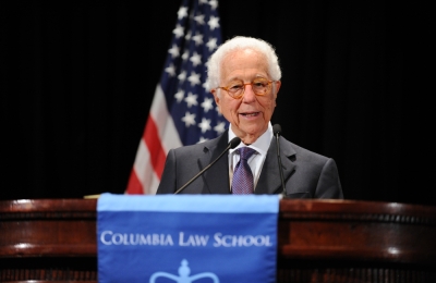 Ira Millstein stands behind a podium and in front of the American flag.