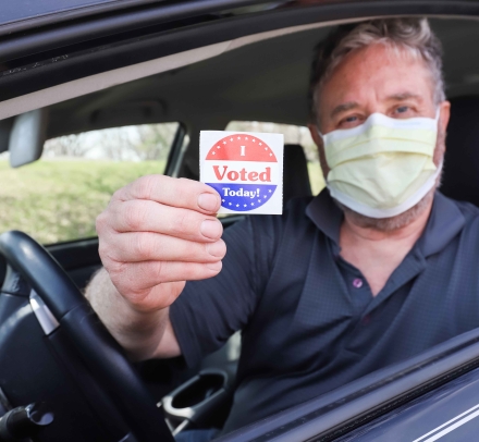 Man wearing a surgical mask for COVID-19 pandemic holds a sticker that says "I VOTED"
