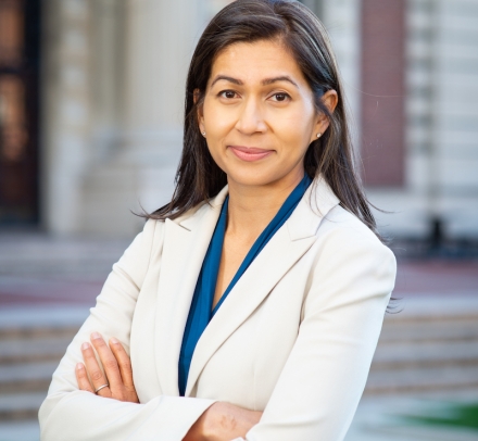 Elora Mukherjee in white jacket and blue blouse with arms crossedd in front of a Columbia University building