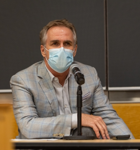 Man in blazer and face mask in front of blackboard