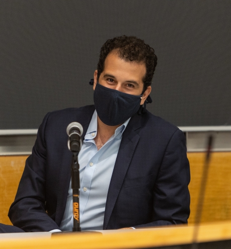 Man in mask wearing light blue shirt and blazer in front of a blackboard