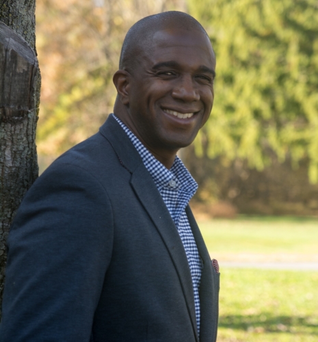 Smiling man in a blue checkered shirt and blue sport coat next to a tree 