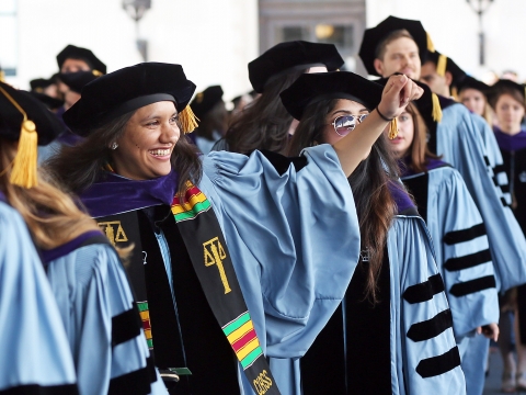 A student wearing academic regalia waves from the processional at the 2017 Graduation.
