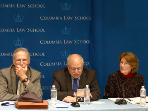 Three people sit at a table with microphones and water bottles in front of them.