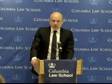 A man in a black blazer standing at a podium in front of a Columbia Law School step and repeat