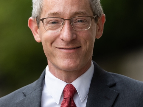 Professor Josh Gupta-Kagan, wearing glasses, a grey jacket, and a red necktie, smiles.