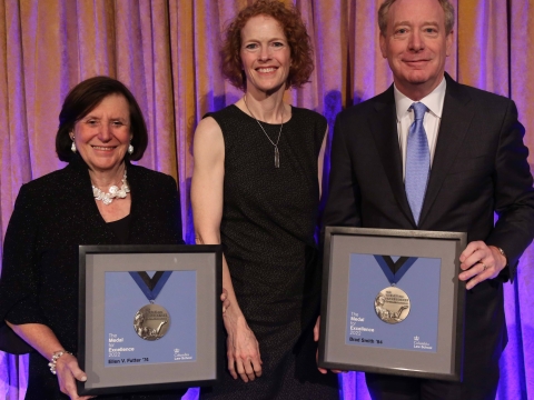 A woman in black flanked by a woman and man, also in black, both holding framed medals.