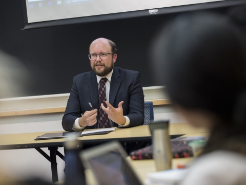 Man in glasses and suit at table in front of a classroom