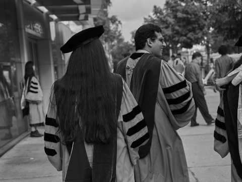 Black and white photo of graduates in gown, scarf, and tam from behind, walking on sidewalk