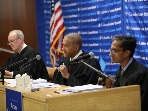 Three men in judges' robes seated behind a desk and speaking into microphones 