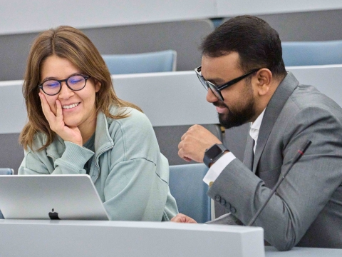 Two smiling students work at a laptop computer in a lecture hall.