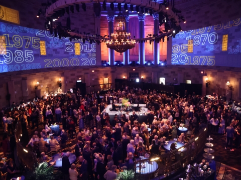Wide shot of room filled with people at a cocktail party with Columbia logo projections on the walls. 