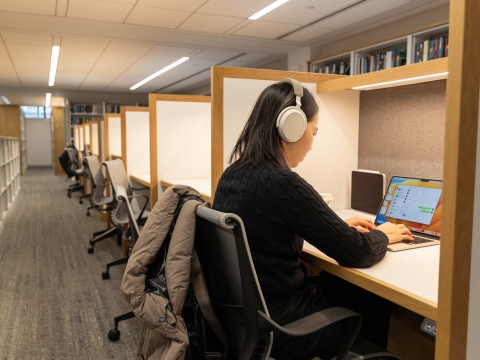 A student wears headphones while studying on a laptop in the library.